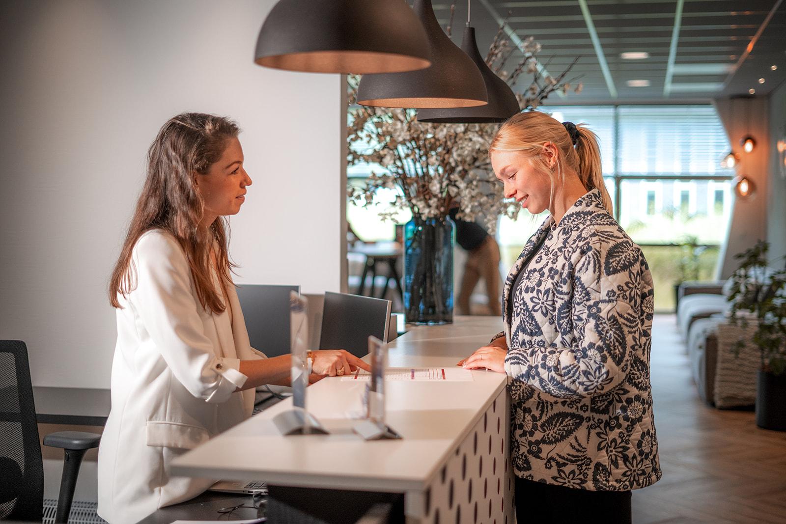 Twee personen bij een moderne witte receptiebalie, één in een wit jasje achter de balie en één in een bloemenprint jasje ervoor, onder stijlvolle zwarte hanglampen met decoratieve bloemenvazen op de achtergrond.